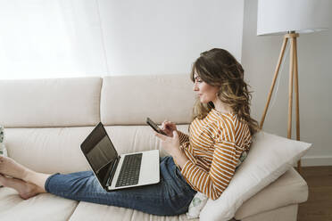 Young woman using laptop and mobile phone on sofa - EBBF06402