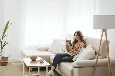 Smiling young woman using digital tablet on sofa at home - EBBF06404