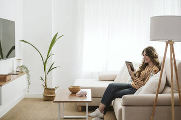 Smiling young woman using digital tablet on sofa at home - EBBF06405