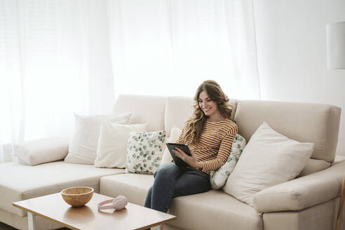 Lächelnde junge Frau, die auf dem Sofa zu Hause ein digitales Tablet benutzt - EBBF06406