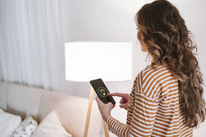 Junge Frau, die ein Mobiltelefon mit einer Smart-Home-App für die Beleuchtung zu Hause benutzt - EBBF06422