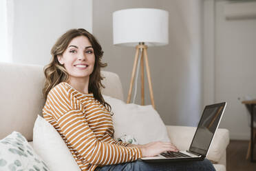 Smiling young woman using laptop on sofa at home - EBBF06425