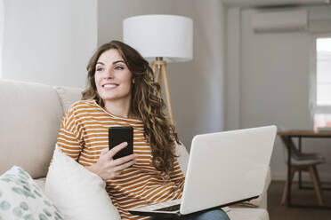 Smiling young woman with laptop and mobile phone on sofa at home - EBBF06426