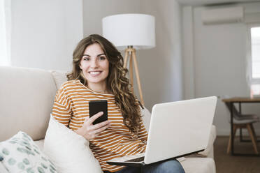 Smiling young woman with laptop and mobile phone on sofa at home - EBBF06427