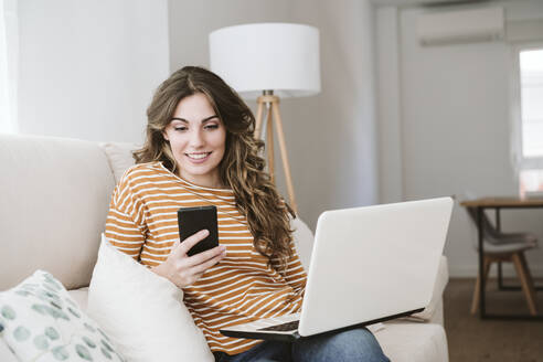 Lächelnde junge Frau mit Laptop und Mobiltelefon auf dem Sofa zu Hause - EBBF06428