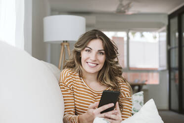 Porträt einer lächelnden jungen Frau mit Mobiltelefon auf dem Sofa zu Hause - EBBF06431