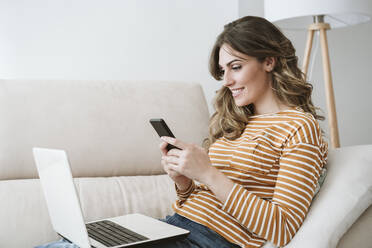 Lächelnde junge Frau mit Laptop und Mobiltelefon auf dem Sofa zu Hause - EBBF06436