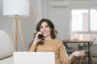 Young woman with laptop on the phone on sofa at home - EBBF06437