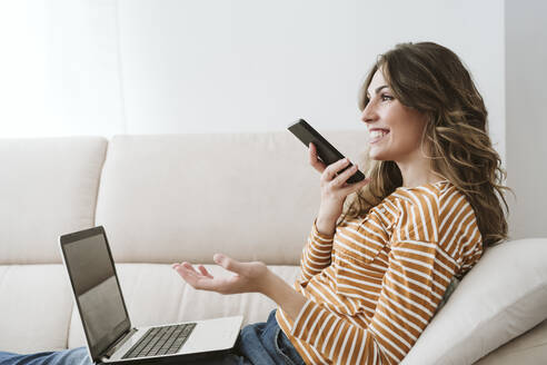 Junge Frau mit Laptop, die zu Hause auf dem Sofa ihr Handy für eine Voicemail benutzt - EBBF06439