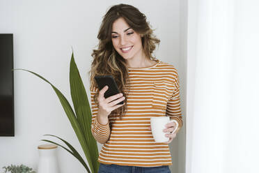 Smiling young woman holding coffee cup and using mobile phone - EBBF06443