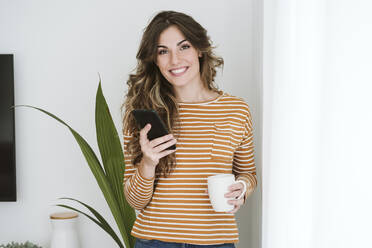 Porträt einer lächelnden jungen Frau mit Kaffeetasse und Mobiltelefon - EBBF06444