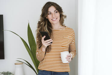 Smiling young woman holding coffee cup and mobile phone - EBBF06445