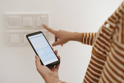 Woman touching switch for blinds while checking mobile phone app - EBBF06453