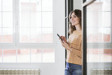 Smiling young woman holding mobile phone at a glass wall at home - EBBF06456