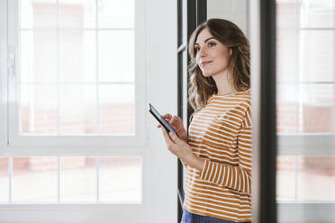 Young woman holding mobile phone at a glass wall at home - EBBF06458