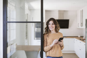 Smiling young woman holding mobile phone at a glass wall in kitchen - EBBF06459