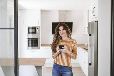 Smiling young woman holding mobile phone in kitchen at home - EBBF06462