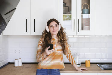 Young woman listening to music with headphones in kitchen at home - EBBF06466