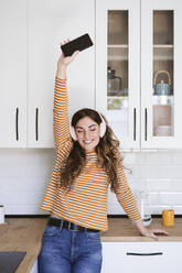 Cheerful young woman listening to music with headphones in kitchen at home - EBBF06467