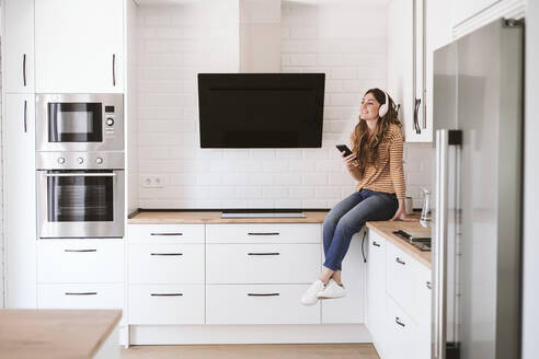 Young woman listening to music with headphones in kitchen at home - EBBF06469