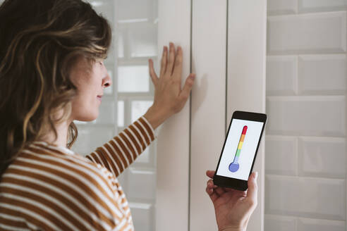 Young woman using smart phone app and touching radiator at home - EBBF06475