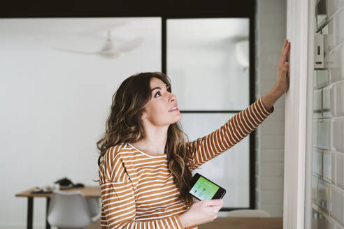 Young woman using smart phone app and touching radiator at home - EBBF06480