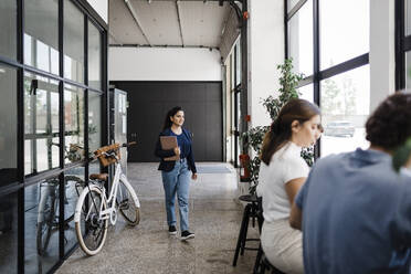 Junge Geschäftsfrau mit Laptop, die mit dem Fahrrad im Flur eines Büros spazieren geht - DCRF01349