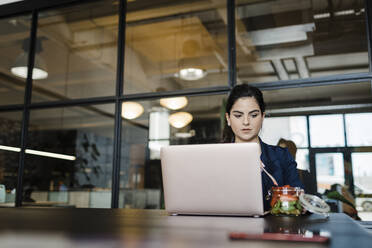 Junge Frau mit Früchten am Laptop im Büro - DCRF01381