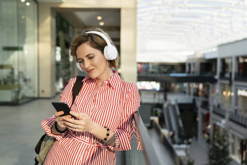 Lächelnde Frau hört Musik über drahtlose Kopfhörer mit einem Smartphone in einem Einkaufszentrum - EKGF00105