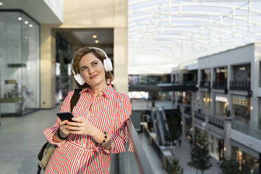 Smiling thoughtful woman listening music through wireless headphones holding smart phone in shopping mall - EKGF00106