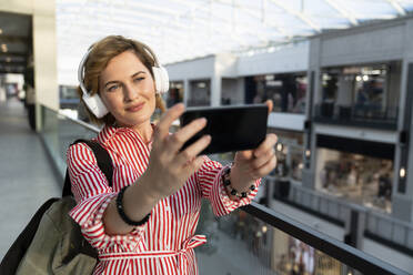 Lächelnde Frau mit drahtlosen Kopfhörern, die ein Selfie mit ihrem Mobiltelefon in einem Einkaufszentrum macht - EKGF00107