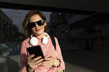 Smiling woman wearing sunglasses using smart phone in shopping mall - EKGF00109