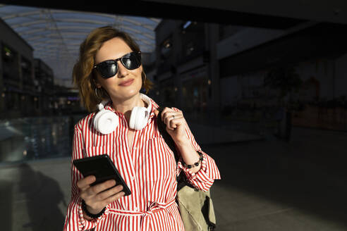 Lächelnde Frau mit Sonnenbrille, die ein Mobiltelefon in einem Einkaufszentrum hält - EKGF00110