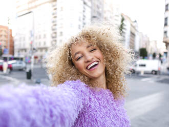 Happy woman with blond Afro hairstyle taking selfie in city - AMRF00046