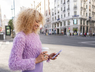 Lächelnde Frau mit Einweg-Kaffeebecher und Smartphone in der Stadt - AMRF00054