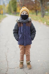 Boy standing with hands in pockets wearing protective face mask on footpath - ONAF00024