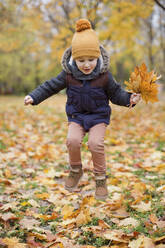 Playful boy holding autumn leaves jumping in park - ONAF00026