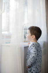 Boy wearing pajamas looking through window at home - ONAF00072