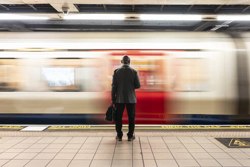 Senior passenger with laptop bag standing in front of speeding train on subway platform - WPEF06403