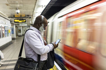 Glatzköpfiger älterer Geschäftsmann benutzt Smartphone in der Nähe eines rasenden Zuges am Bahnhof - WPEF06410