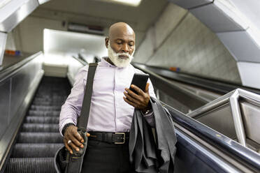 Senior commuter with laptop bag using smart phone on escalator - WPEF06411