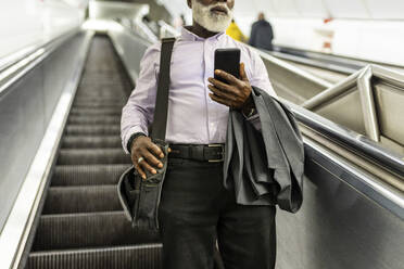 Älterer Geschäftsmann mit Anzug und Mobiltelefon, der eine Laptoptasche auf einer Rolltreppe trägt - WPEF06412