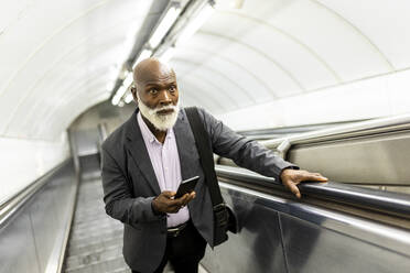 Älterer Geschäftsmann mit Smartphone, der auf einer Rolltreppe nach oben fährt - WPEF06414