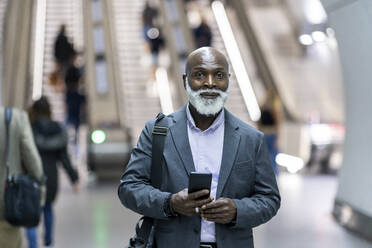 Smiling senior businessman with laptop bag and mobile phone at railroad station - WPEF06424