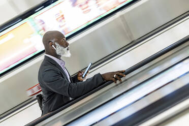 Älterer Geschäftsmann mit Smartphone, der auf einer Rolltreppe in einer U-Bahn-Station nach oben fährt - WPEF06439