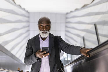 Älterer Pendler mit Smartphone, der auf einer Rolltreppe im Bahnhof nach oben fährt - WPEF06446