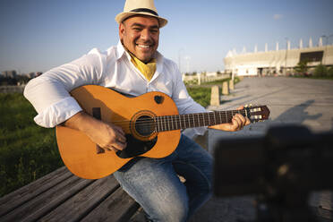 Lächelnder Gitarrist spielt Gitarre und nimmt mit seinem Smartphone auf einem Stativ auf - EKGF00113
