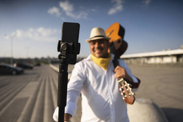 Lächelnder Gitarrist, der eine Gitarre hält und ein Selfie mit seinem Mobiltelefon auf einem Stativ macht - EKGF00118