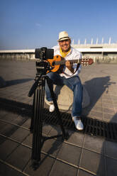 Smiling street musician playing guitar and recording through smart phone on tripod - EKGF00124