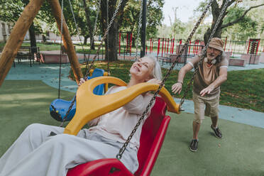 Senior man pushing woman enjoying swing at park - YTF00042